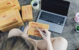 mulher sentada no chão em frente a um notebook, preenchendo endereços em caixas que está vendendo na internet