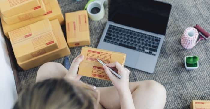 mulher sentada no chão em frente a um notebook, preenchendo endereços em caixas que está vendendo na internet