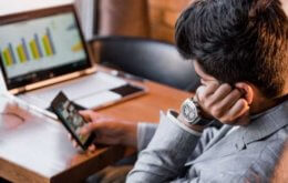 homem entediado olhando para o celular enquanto tem trabalho para fazer, representando o conceito de vencer a procrastinação